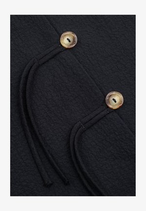 Zbliżenie czarnego teksturowanego materiału z dwoma beżowymi, okrągłymi guzikami oraz przeszywanymi, zakrzywionymi ozdobnymi pętlami.