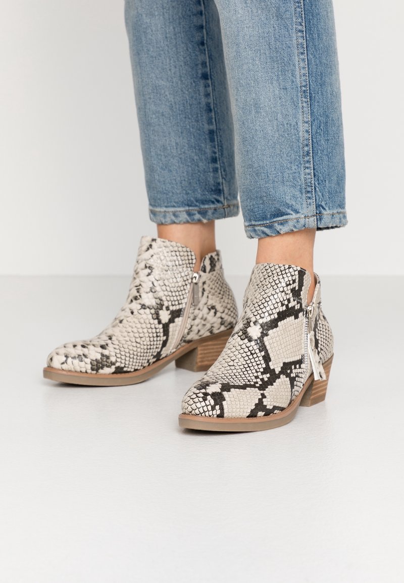 Bottines avec motif serpent beige et noir, fermeture éclair latérale, petit talon en bois, portées avec un jean bleu retroussé sur un sol blanc.