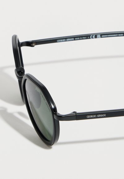 Μαύρα στρογγυλά γυαλιά ηλίου Giorgio Armani με λεπτή σκελετό και σκοτεινούς φακούς πάνω σε λευκή επιφάνεια.