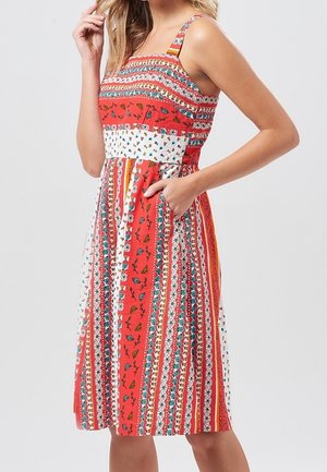 Kobieta ubrana w sukienkę bez rękawów do kolan z czerwonymi, białymi i wielokolorowymi pionowymi paskami oraz kwiatowymi wzorami, z jedną ręką w kieszeni.