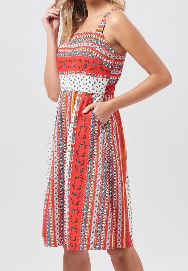Femme portant une robe sans manches, longueur genou, avec des rayures verticales rouges, blanches et multicolores ainsi que des motifs floraux, avec une main dans la poche.