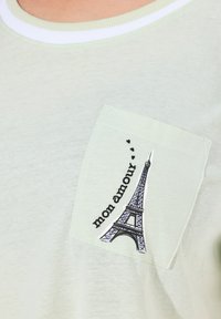 Ljusgrön t-shirt med en vit ribbad halsringning. Har en ficka med en svart skiss av Eiffeltornet och texten "mon amour."