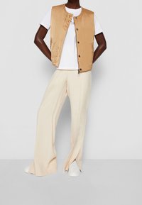 Tan satin vest over a white t-shirt, paired with cream wide-leg pants and white footwear. Smooth texture, minimal buttons, and casual design.