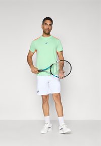 Bărbat îmbrăcat într-o cămașă sport verde și portocalie și pantaloni scurți albi, ținând o rachetă de tenis, stând în fața unui fundal simplu, deschis la culoare.