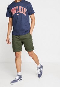 Laivaston t-paita, jossa on punaisella tekstillä "TOMMY JEANS", oliivinvihreät shortsit, valkoiset sukat ja laivastonväriset lenkkarit valkoisilla yksityiskohdilla.