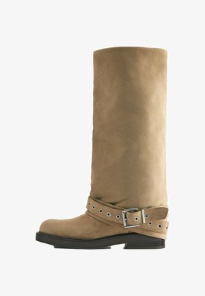 Botte haute jusqu'au genou en daim beige avec semelle noire et bride décorative avec boucle argentée autour de la cheville.
