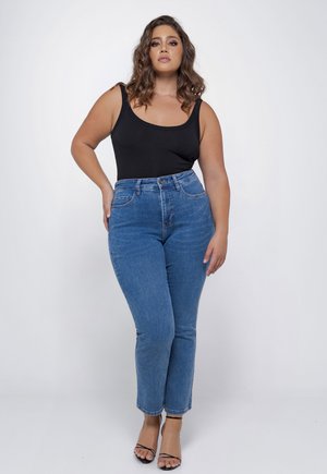 Kroeshaar vrouw draagt een zwarte mouwloze top, hoge taille blauwe jeans en zwarte sandaalhakken, staande tegen een witte achtergrond.