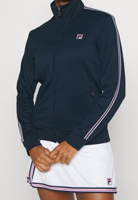 Námořnická zipová bunda s vysokým límcem, doplněná bílými a červenými bočními pruhy. Vyrobena z hladké látky s logem na hrudi.