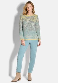 Vrouw in pastelblauwe broek, tan enkelboeksen en een langemouwen, kleurrijke gebreide truien staande tegen een witte achtergrond.
