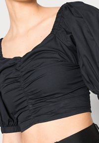 Top negro corto con escote cuadrado, mangas abullonadas y detalles fruncidos en la parte delantera y los laterales. Textura de tela suave y lisa.