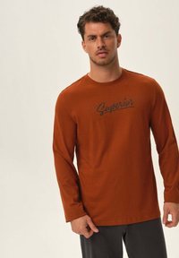 Długo rękawowa, rdzawa koszula z miękkiego materiału, z czarnym nadrukiem logo "Superior" na piersiach, z okrągłym dekoltem.