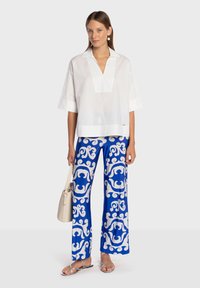 Chemise blanche à col avec encolure en V, associée à un pantalon large bleu orné d'un motif blanc tourbillonnant. Le mannequin tient un sac à main clair.