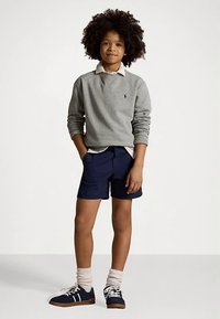 Kihiline laps seisab hallis dressipluusis, valges särkis, tumesinistes lühikeses jaos, valgete sokkide ja tumesiniste tossudega valge taustaga.