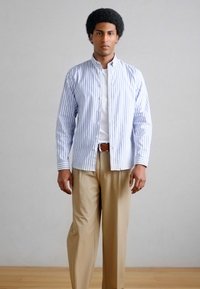 Άνδρας όρθιος, στραμμένος προς τα εμπρός, φορούν μπεζ παντελόνι, λευκό μπλουζάκι και πουκάμισο με κάθετες ρίγες μπλε και λευκό.