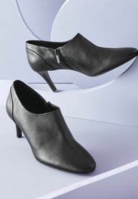 Musta nahkabootit, joissa on terävät varpaat, sivussa vetoketjut ja 3-tuumaiset stiletto-korkokengät, esillä vaalean violetilla taustalla.