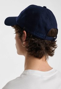 Person med lockigt brunt hår som bär en marinblå keps och vit skjorta, visad från bakom och vänd mot vänster.