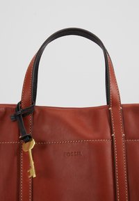 Braune Leder-Tote-Tasche mit schwarzen Akzenten, ausgestattet mit einem schlüsselförmigen goldenen Anhänger und sichtbaren Nähten, auf dem der Markenname "FOSSIL" zu sehen ist.