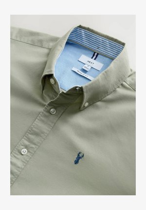 Kerge oliivroheline nööbiga särk, mis on valmistatud siledast kangast, sinise triibulise krae voodriga ja väikese tumesinise tikandiga.