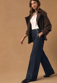 Donkerblauwe jeans met hoge taille en wijde pijpen, met gouden knoopaccenten, gecombineerd met een wit shirt en een bruine jas, tegen een neutrale achtergrond.