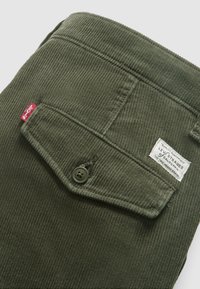 Olivgrüner Cordstoff mit einer Knopfklapptasche, ausgestattet mit einem weißen Etikett mit Markendetails. Strukturierte Oberfläche, gerade Form.