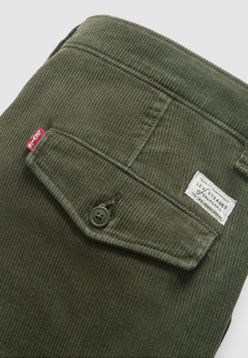 Olive green corduroy fabric with a buttoned flap pocket, featuring a white label with brand details. Textured surface, straight shape.