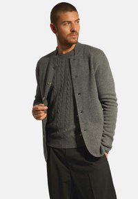 Šedý cardigan s kulatým výstřihem, zapínáním na knoflíky a ribbovanými manžetami, přes tmavě šedý svetr s kabelkovým pletením. Hladká, texturovaná látka.