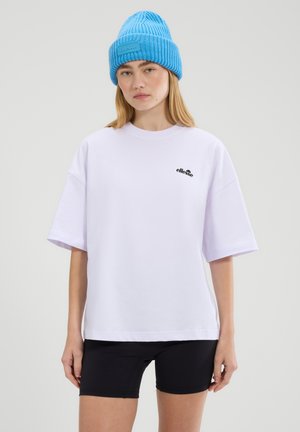 Young woman wearing a light blue knit beanie, white oversized t-shirt with small logo, and black fitted shorts, standing against a plain background.