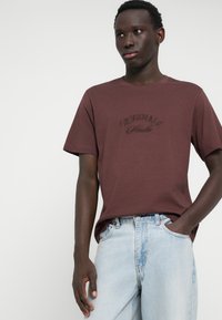 Bordeauxkleurig t-shirt met korte mouwen en reliëftekst "Originals", gecombineerd met lichtblauwe spijkerbroek. Glad materiaal en relaxed fit.