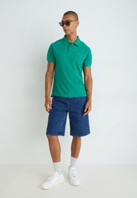 Zelené polo tričko, modré denimové šortky, biele tenisky, šedé ponožky, doplnené slnečnými okuliarmi. Krátke rukávy a klasický dizajn goliera.