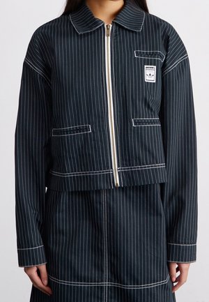 Frau trägt eine schwarze Nadelstreifenjacke mit Reißverschluss, weißer Naht und einem Adidas-Logo auf der Brust, kombiniert mit einem passenden Rock.