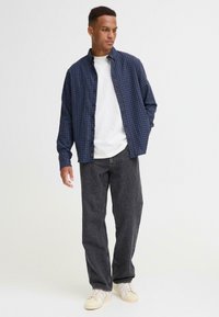 Geruite shirt in marineblauw en rood, witte t-shirt eronder, grijze denim jeans en lichtgekleurde sneakers, staande op een neutrale achtergrond.