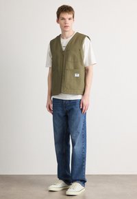Gilet sans manches vert olive avec une fermeture éclair à l'avant, deux poches poitrine, porté sur un t-shirt blanc et un jean bleu, assorti à des baskets crème.