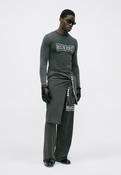 Mann mit schwarzen Sonnenbrillen, langärmligem Grafikshirt, einem um die Taille gebundenen Sweatshirt, weiten Hosen, schwarzen Handschuhen und schwarzen Schuhen, der vor einem weißen Hintergrund steht.