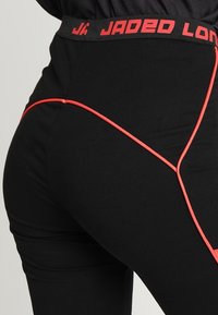 Černé legíny s červeným elastickým pasem označeným "JADED" a červeným lemováním po stranách. Hladká textura, přiléhavý střih.