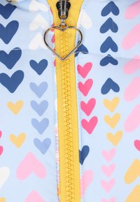 Polstret jakke med en lyseblå baggrund og farverige hjerte mønstre i pink, gul og marineblå samt en gul lynlås med hjerte-formet træk.