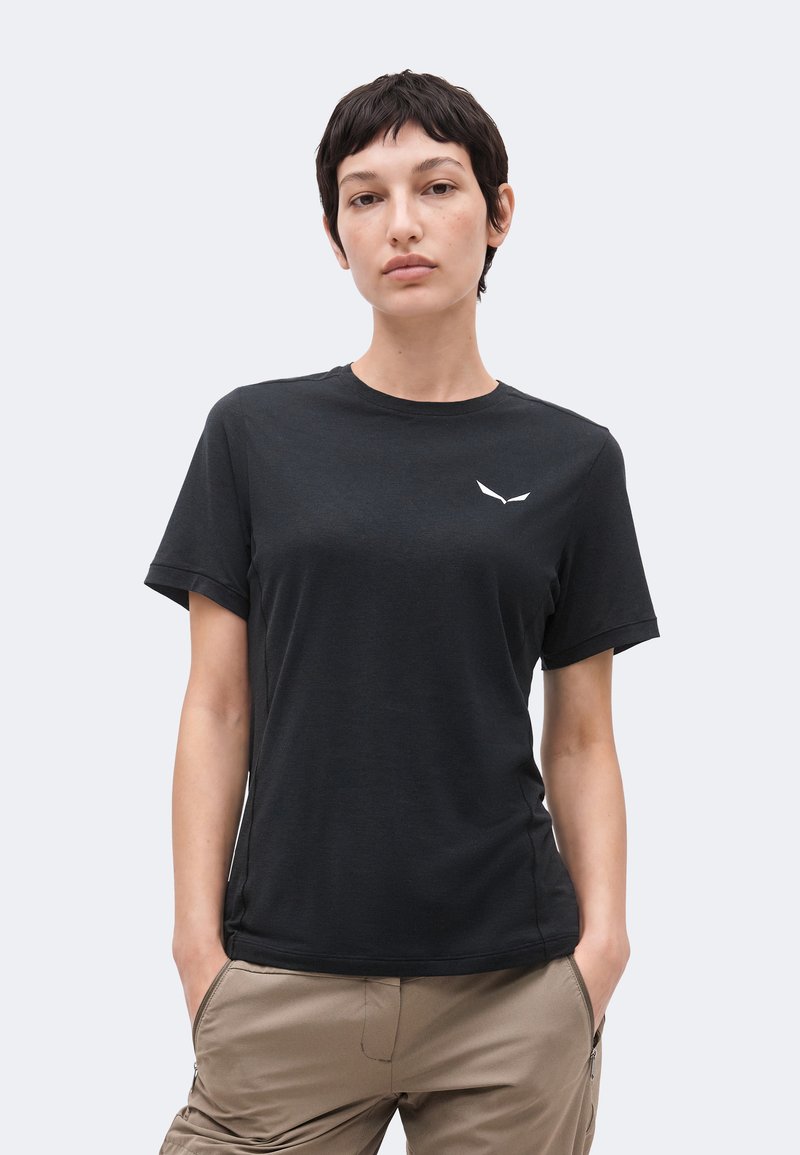 Νεαρό άτομο με κοντά σκούρα μαλλιά που φοράει μαύρο t-shirt με στρογγυλή λαιμόκοψη και μικρό λευκό λογότυπο στο στήθος, χέρια στις τσέπες μπεζ παντελονιού.