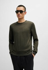 Olivegrüner Pullover mit langen Ärmeln und Rundhalsausschnitt, aus einem strukturierten Material gefertigt. Kombiniert mit schwarzen Sonnenbrillen und dunklen Hosen.