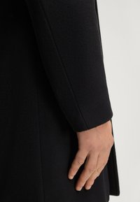 Zwarte jas van gestructureerde stof. Heeft een getailleerde pasvorm met een rechte mouw en subtiele naaddetails. Close-up van de mouw en hand.