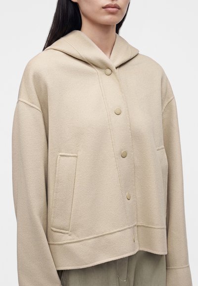 Femme portant une veste à capuche en laine beige avec des boutons-pression à l'avant et des poches latérales, visible des épaules à la taille.