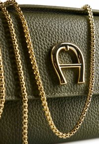 Bolso de cuero verde texturizado con una correa de cadena metálica dorada y un destacado logo dorado en la parte frontal.