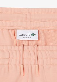 Lichtroze elastische tailleband met bijpassende trekkoorden en een wit label met daarop "Lacoste" en "Regular Fit" met een groen krokodillenlogo.