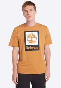 Camiseta de algodón marrón con un gran gráfico cuadrado blanco que presenta un diseño de árbol y el texto "Timberland" en negro debajo. Mangas cortas.