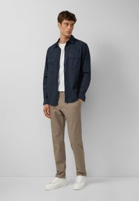 Navy-blaues Hemd mit zwei Brusttaschen, kombiniert mit beigen Hosen und weißen Sneakers, vor einem hellgrauen Hintergrund stehend.