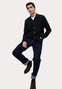 Helleblauer Button-Down-Cardigan aus weichem Material mit langen Ärmeln, kombiniert mit dunkelblauen Jeans, hochgekrempelten Ärmeln und braunen Schnürschuhen.