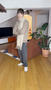 Beige knit sweater with a cream scarf, gray jeans, and white sneakers. Standing in a room with a brown corduroy sofa and green plant.