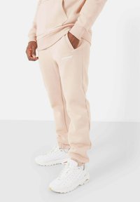Lichtroze sweatpants van zacht materiaal, met elastische boorden, zijzakken en een klein wit logo op de linker dij. Gecombineerd met witte sneakers.