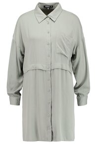 Ljusgrå skjorta med knappar, långärmad, med krage och en enda bröstficka; har en insnörning i midjan och en mjuk textur.