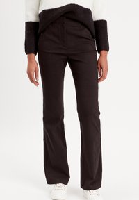 Personne portant un pantalon noir taille haute évasé, un pull en tricot noir et blanc, et des baskets blanches, se tenant devant un fond blanc uni.
