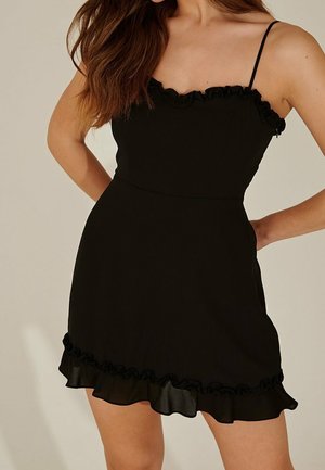 Mujer con un vestido mini negro sin mangas, con tirantes finos y ribete con volantes en el escote y el dobladillo, de pie con las manos en las caderas.