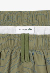 Olijfgroene zwembroek met blauw contourpatroon, elastische tailleband, witte meshvoering en een Lacoste logo-label. Voorzien van een verstelbaar trekkoord.